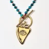 Χειροποίητο κολιέ σε χρυσό χρώμα με σχέδιο καρδιά με σκαλιστό μάτι και αλυσίδα από γαλαζοπράσινους ημιπολύτιμους λίθους. Διάσταση 43εκ. Σχεδιάστρια Νεφέλη Καρυοφύλλη.